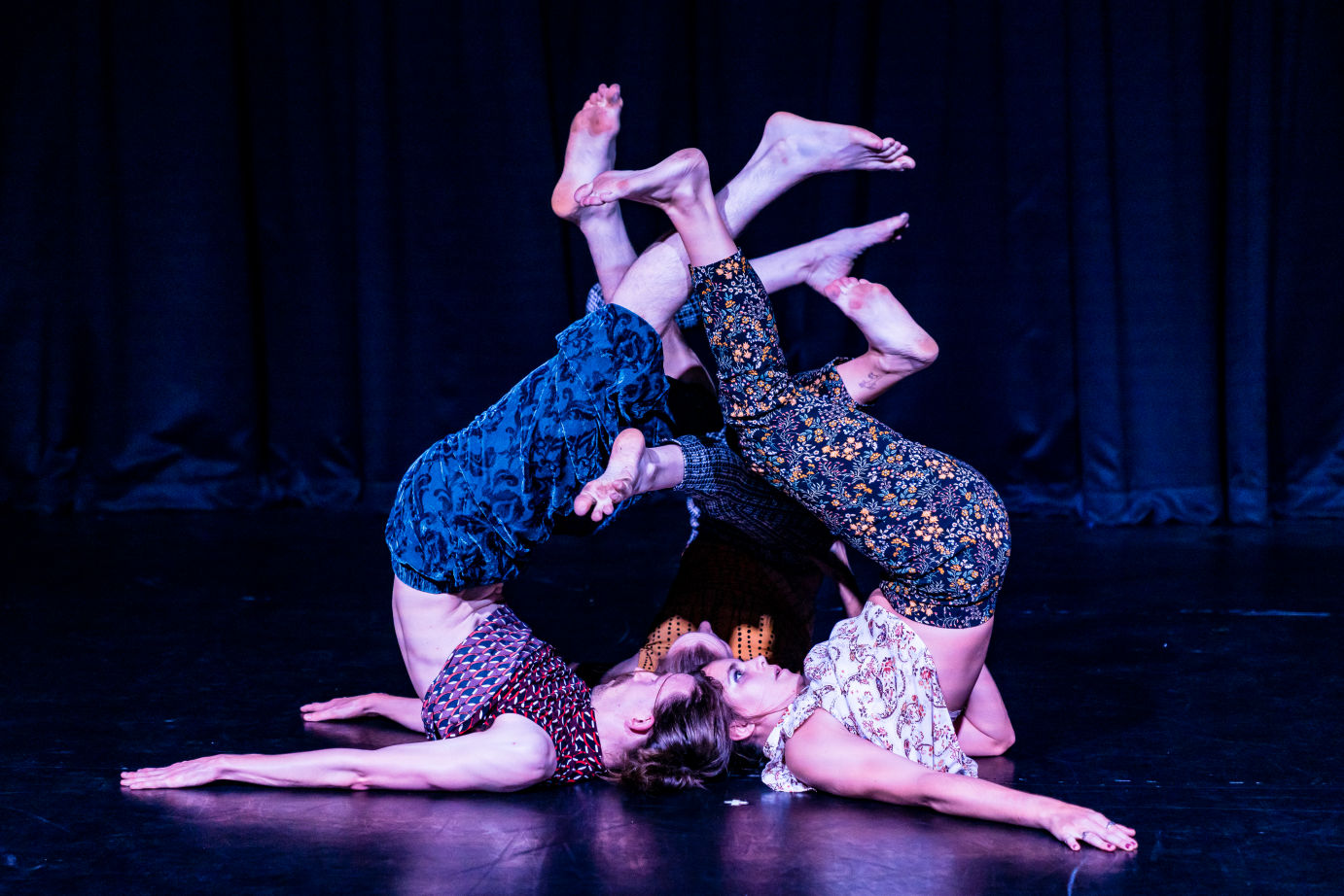 In a clump, three people do shoulder stands on the floor. Their feet entwine.